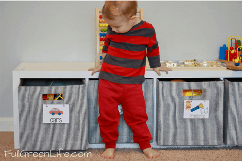 2-year-old boy standing in front of toy bins that are labeled with words and pictures- "cars" and "pretend"