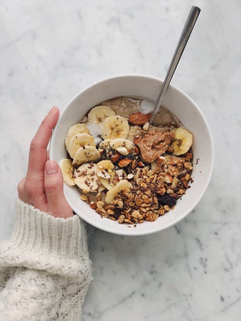 woman's hand holding a breakfast bowl filled with meusli, nuts, bananas, and dried fruit, with a spoon
