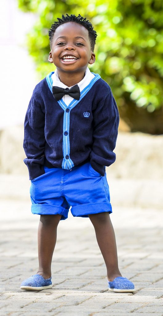 little boy wearing shorts, a cardigan and a bow tie with his hands in his pockets, smiling