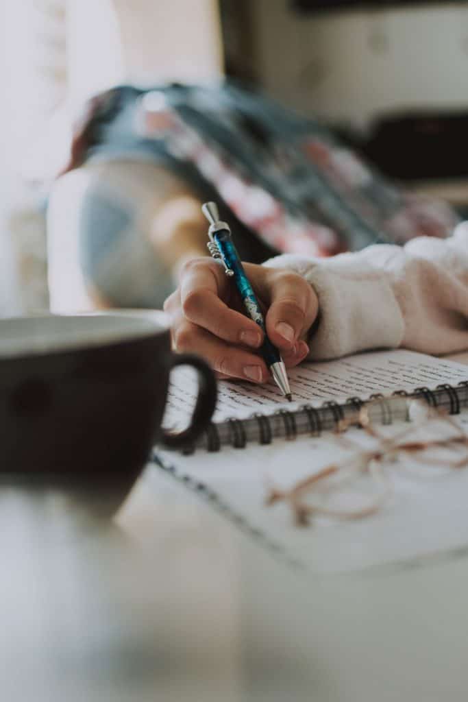 closeup of woman holding a blue pen writing in a journal, black coffee cup in foreground